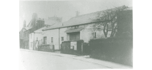 The barn in good condition, a good wall in front, double doors closed. The church tower just seen above the roofs in the background behind the barn and adjacent buildings