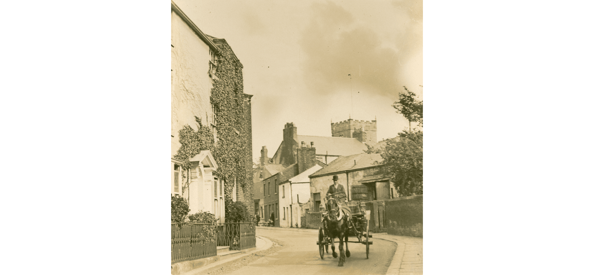 Tithebarn Street, an old sepia photo. We see the church tower behind. In the foreground a horse and cart is being driven towards us by a man in a bowler hat. Ivy covers the front of the houses on our left
