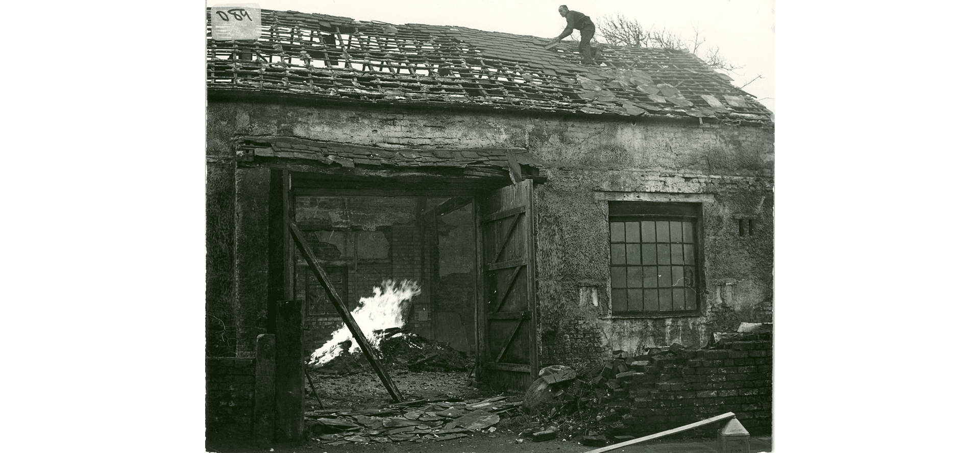 The tithebarn in Poulton being demolished. A man on the roof removing tiles. A fire can be seen through the open double barn doors, inside the building.