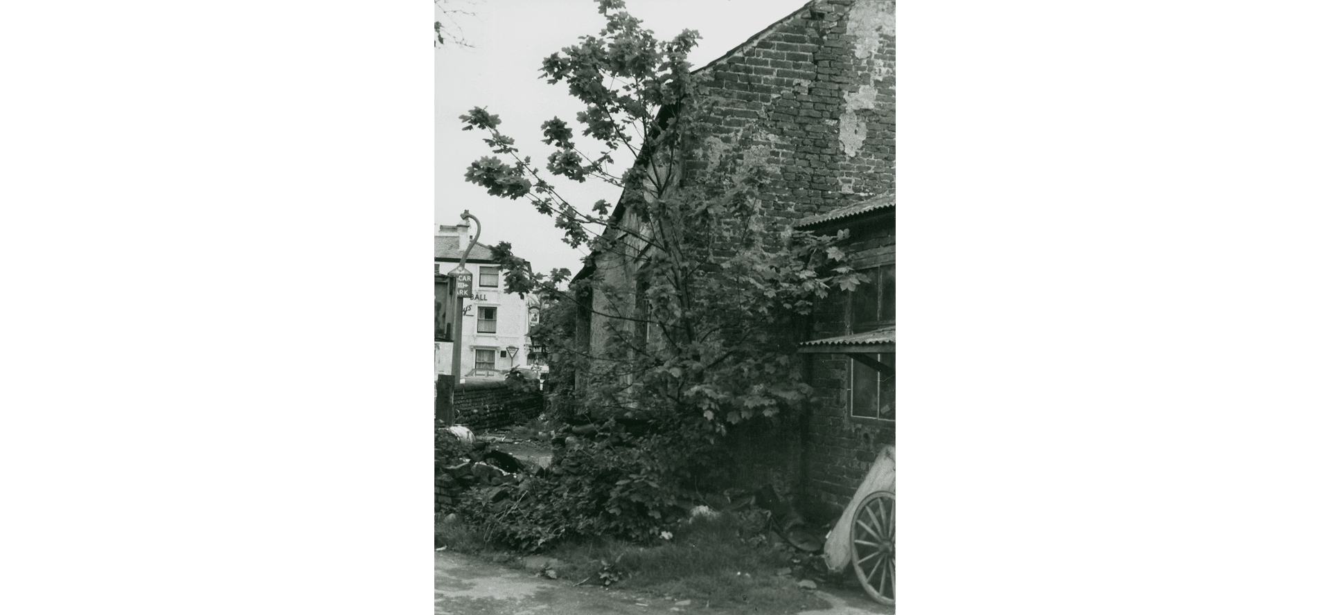 The Tithebarn in Poulton. We see the corner of an old building, a tree growing out of it, and an old wooden wheel in the foreground.