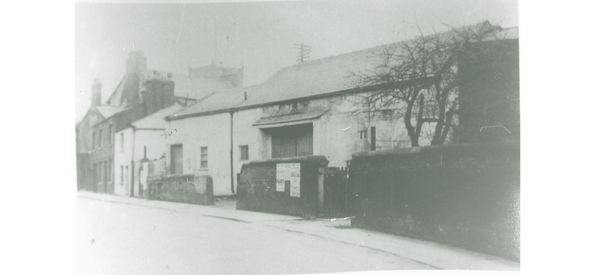 The barn in good condition, a good wall in front, double doors closed. The church tower just seen above the roofs in the background behind the barn and adjacent buildings